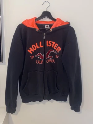 Mörkgrå Hollister hoodie med dragkedja - Hoodie från Hollister med röd-orange huva och broderad logga framtill. Dragkedja hela vägen, två fickor och ribbade muddar. Stora tryckta bokstäver och fågelmotiv ger en sportig vibe. Perfekt för chill dagar eller när du vill ha en casual look.