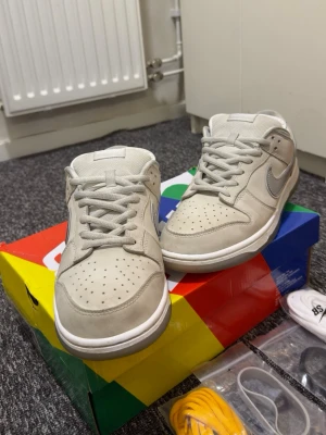 Nike Dunk Low SB grå/vita sneakers - Nike Dunk Low SB sneakers i ljusgrå och vit färg med silvergrå swoosh. Skorna har perforerad tå, platt sula och rund tå. Materialet är skinn och textil, och innersulan har ett rutigt mönster. Flera extra skosnören medföljer i olika färger.