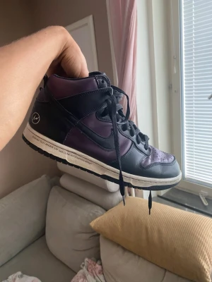Svarta och lila höga sneakers - Säljer ett par höga sneakers i svart och lila med svart snörning och vit sula. Skorna har en klassisk siluett med perforerad tåbox och detaljer i läder. Snygg kontrast mellan färgerna och en ikonisk look som sticker ut.
