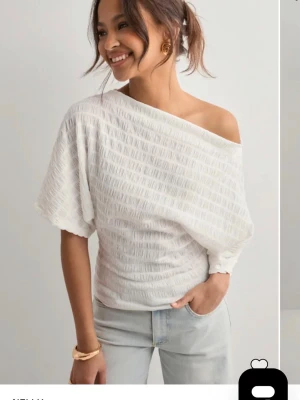 Vit offshoulder topp med struktur - Trendig vit topp med offshoulder-design och strukturerat tyg. Toppen har kort ärm och en lös, avslappnad passform upptill som smiter åt vid midjan. Perfekt för dig som vill ha en unik och stilren look. Den är aldrig använd , köpte den men den passade inte mig och tiden att skicka tillbaka hade gått ut så därför säljer jag den