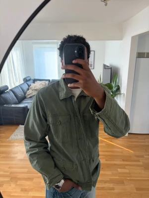 Grön vintage jacka med knappar och dragkedja från kangol  - Snygg militärgrön jacka med fyra fickor framtill och knäppning hela vägen. Jackan har en avslappnad passform och är tillverkad i bomull, perfekt för lager på lager under vår och höst.