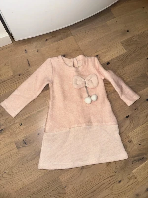 Ljusrosa klänning med rosett och bollar - Supersöt ljusrosa klänning från Lilax med lång ärm och dekorativ rosett framtill samt två fluffiga bollar. Klänningen har en mjuk, mysig känsla och är perfekt för dig som gillar gulliga detaljer.Ljusrosa klänning med rosett och bollar