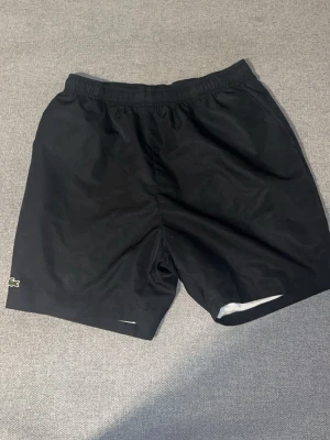 Svarta Lacoste shorts - Säljer ett par svarta shorts med resår i midjan och enkel design. Shortsen har en liten Lacoste  logga på vänster ben och är tillverkade i ett lätt material som passar perfekt till garderoben 