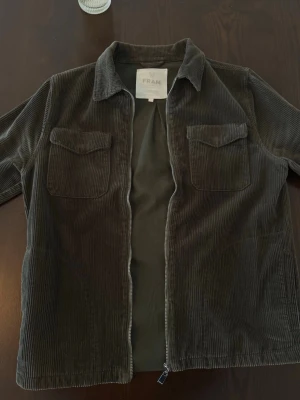 Mörkgrön manchester overshirt FRAM - Snygg mörkgrön overshirt i manchestertyg från FRAM. Jackan har två bröstfickor med lock, klassisk krage och dragkedja framtill. Perfekt lager på lager-plagg med avslappnad passform och mjuk känsla.