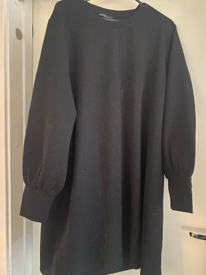 Svart glittrig klänning Mywear Woman - Snygg svart kortklänning från Mywear Woman med diskret glittrigt tyg. Klänningen har långa puffärmar med breda muddar och en rak, avslappnad passform. Perfekt för dig som vill ha något enkelt men ändå lite extra.