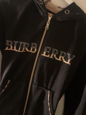 Svart hoodie med dragkedja från Burberry - Snygg svart hoodie från Burberry med dragkedja framtill och huva. Stora Burberry-bokstäver i strass och beige mönster på bröstet samt detaljer vid fickorna. Klassisk Burberry-tag på insidan. Perfekt för en trendig och avslappnad stil.