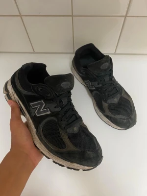  New Balance M2002R Sneakers  - Säljer ett par svarta New Balance sneakers med ovandel i mesh och mocka. Skorna har klassisk N-logga på sidan, svarta skosnören och vit sula med slitna detaljer. Perfekta för en avslappnad streetwear-look.