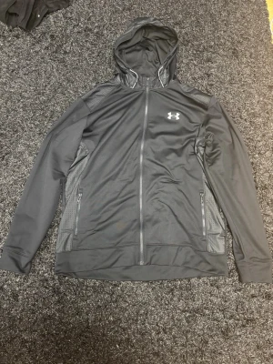 Svart hoodie från Under Armour - Snygg svart hoodie från Under Armour med dragkedja framtill, två fickor med dragkedja och meshdetaljer på axlar och huva. Tillverkad i ett sportigt, tekniskt material som andas. Perfekt för träning eller chill. Under Armour-logga på bröstet och ryggen.