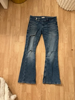 Blå bootcut jeans med slitningar - Säljer ett par blå bootcut jeans med slitningar på knäna och framsidan. Jeansen har klassiska bakfickor och framfickor samt bälteshällor. Materialet är denim med en lätt tvättad look och slitna detaljer för en trendig vibe.