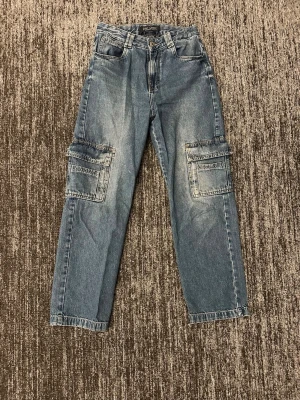 Blå cargobyxor i denim - Säljer ett par blå cargobyxor i klassisk denim med raka ben och stora fickor på sidorna. Byxorna har normal midja, bälteshällor och två bakfickor. Perfekta för en avslappnad och trendig streetwear-look.