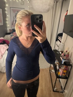 Mörkblå topp med spetsdetalj - Snygg mörkblå långärmad topp med djup v-ringning och spetsdetaljer vid halsen. Toppens material känns mjukt och stretchigt, perfekt för en trendig look. Passformen är normal och toppen är enkel att matcha med olika outfits.