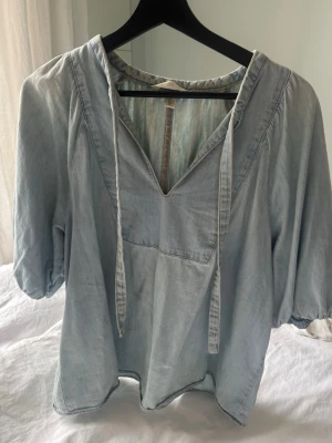 Jeans-topp - Ljusblå blus i jeans-imitation med djup v-ringning och knytband vid halsen. Blusen har trekvartsärm med lätt puff och avslappnad passform. Sparsamt använd! 