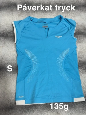 Blå sportig topp från Reebok - Sportig blå topp från Reebok med vita detaljer och korta ärmar. Toppen har ett diskret mönster på sidorna och liten v-ringning framtill. Perfekt för dig som gillar en aktiv stil.🌸 påverkat tryck. Storlek s