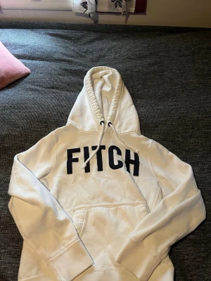 Vit hoodie från Abercrombie & Fitch - Snygg vit hoodie från Abercrombie & Fitch med stor svart text 'FITCH' på bröstet. Klassisk känguruficka framtill, ribbade muddar och justerbar huva med snören. Mjuk insida för extra komfort, perfekt för chill dagar. Säljer pga att den var för liten, skickas dagen den beställs eller dagen där efter!