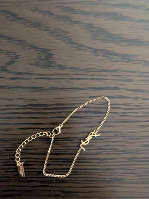 Guldfärgat armband från Saint Laurent - Snyggt och stilrent armband i guldfärgad metall från Saint Laurent. Armbandet har en tunn kedja och ikoniska YSL-loggan i mitten. Justerbar längd med liten bricka där märket står ingraverat. Perfekt accessoar för att lyfta din outfit.