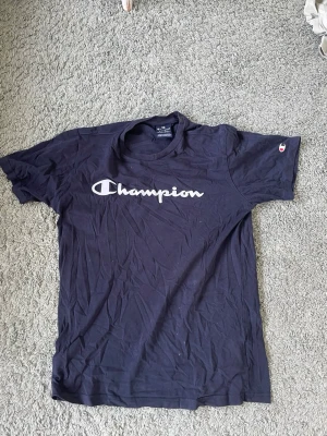 Mörkblå Champion t-shirt - Snygg mörkblå t-shirt från Champion med klassisk logga tryckt i vitt över bröstet och liten logga på ärmen. T-shirten har rund hals och korta ärmar, perfekt för en avslappnad och sportig stil.