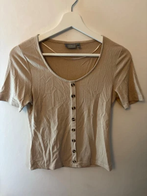 Beige ribbad topp med knappar - Snygg beige ribbad topp med korta ärmar och knappar framtill. Toppen har en rundad urringning och är tillverkad i ett mjukt, stretchigt material som sitter bekvämt. 