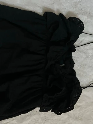 Svart kort linne med volang - Söt svart kort linne med volangdetaljer och tunna axelband. Linnet har broderade hålmönster och en luftig passform som ger en romantisk känsla. Perfekt för dig som gillar en feminin och trendig stil.