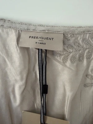 Silverfärgad klänning från Freequent - Snygg silverfärgad klänning från Freequent i storlek XL. Klänningen har v-ringning och är dekorerad med broderade blommor i matchande ton. Materialet har en skimrande finish som ger en exklusiv känsla. Perfekt för dig som vill sticka ut.