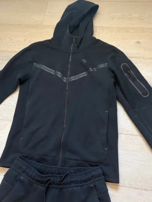 Nike tech fleece dress - Säljer en svart mjukisdress med dragkedja och huva. Tröjan har en snygg detalj med reflexband över bröstet och en ficka med dragkedja på ärmen. Byxorna har resår i midjan och dragsko för skön passform. Perfekt för chill dagar hemma.