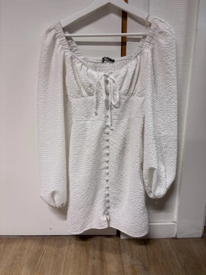 Vit klänning från Gina Tricot - Supersöt vit klänning från Gina Tricot med puffiga långärmar, knytning vid bysten och dekorativa knappar framtill. Klänningen har en strukturerad yta och offshoulder-modell som ger en trendig vibe. Bra skick i stl 34. Hör av dig vid fler bilder eller andra frågor!💕