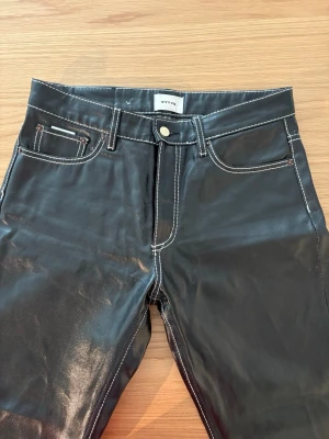 Svarta coated jeans från Eytys - Säljer ett par svarta coated jeans från Eytys med raka ben och kontrastsömmar i vitt. Jeansen har klassisk femficksmodell och metallknapp i midjan. Materialet har en glansig finish som ger en cool look.