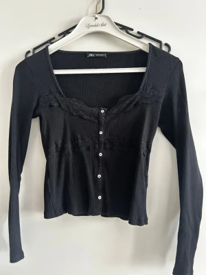 Svart ribbad topp med spets från Zara - Trendig svart ribbad topp från Zara med långa ärmar och knappar framtill. Toppen har fina spetsdetaljer längs ringningen och under bysten, vilket ger en feminin touch. Perfekt till jeans eller kjol för en snygg look. Använd fåtal gånger, har ett mindre hål under spetsen där sömmen har lossnat från tyget - går att laga.