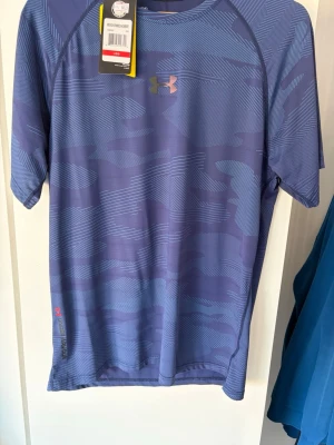 Blå tränings-tshirt från Under Armour - Använd 1 gång. Snygg blå tränings-tshirt från Under Armour med diskret mönster i olika blå nyanser. T-shirten har korta ärmar, rund halsringning och logga på bröstet samt vid nederkanten. Tillverkad i ett lätt och snabbtorkande funktionsmaterial.
