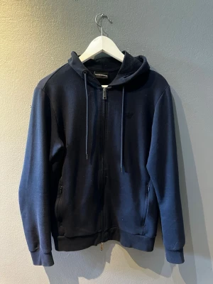 Mörkblå hoodie från Emporio Armani - Snygg mörkblå hoodie från Emporio Armani med dragkedja framtill, två fickor och justerbar huva med snören. Tillverkad i mjukt bomullsmaterial och har diskret logga broderad på bröstet. Perfekt för en avslappnad och stilren look.