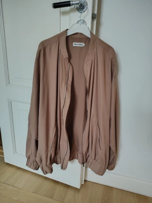 Beige bomberjacka från Ahlvar Gallery - Säljer en snygg beige-rosa bomberjacka från Ahlvar Gallery. Jackan har en avslappnad passform, dragkedja framtill, fickor på sidorna och resår vid ärmslut och nederkant. Tillverkad i ett mjukt, tunt material som ger en stilren look.