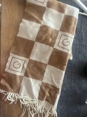 Beige och vit rutig halsduk från Gant - Snygg halsduk från Gant i beige och vitt rutmönster med fransar i ändarna. Tillverkad i mjukt material och har diskreta logodetaljer invävda i rutorna. Perfekt accessoar för att lyfta din outfit med en klassisk touch.