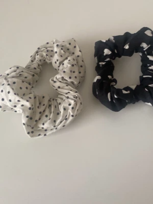 Två scrunchies med mönster - Säljer två scrunchies i tyg, en vit med svarta prickar och en svart med vita hjärtan. Perfekta för att piffa till din frisyr och ge en söt touch till din look. Båda är mjuka och stretchiga, passar de flesta hårtyper.