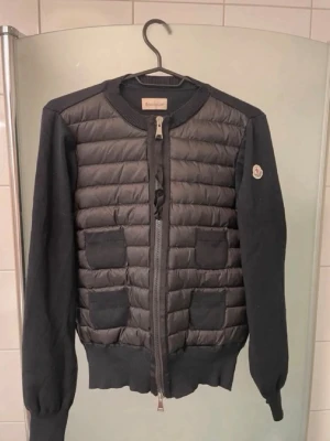 Svart cardiganjacka från Moncler - Snygg svart cardiganjacka från Moncler med quiltad front och stickade ärmar. Jackan har rund halsringning, dragkedja framtill och fyra dekorativa fickor på framsidan. Moncler-logga på ena ärmen. Perfekt för lager på lager och streetstyle.