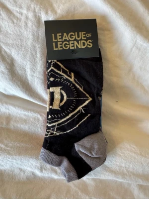 League of Legends strumpor 3-pack - Säljer ett 3-pack League of Legends strumpor i svart, rött och blått med olika tryck och detaljer. Strumporna är låga och har kontrastfärgade tår och hälar. Perfekt för dig som gillar gaming och vill ha något unikt på fötterna.