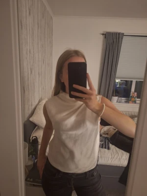 Vit ärmlös satinlinnetopp - Snygg vit ärmlös linnetopp i satin med hög hals och avslappnad passform. Toppen har en lätt glansig yta som ger en lyxig känsla och passar perfekt till jeans eller kjol. Enkel men stilren design som är lätt att matcha. Finns en jätteliten sminkfläck vid nacken.