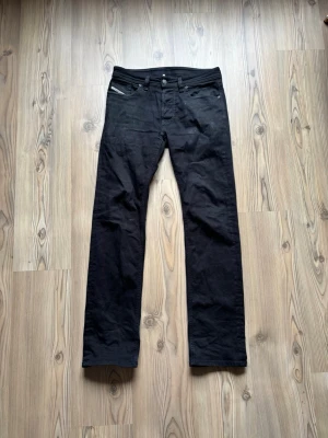 Svarta raka jeans från Diesel - Säljer ett par svarta jeans med klassisk rak passform och fem fickor. Jeansen har bälteshällor, knappgylf och detaljer i silver. Materialet är denim och färgen är helsvart, vilket gör dem enkla att matcha till olika stilar. Storlek 29/32