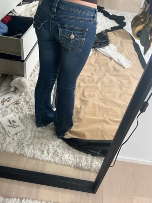 True religion Mörkblå bootcut jeans med detaljer - Säljer ett par mörkblå jeans med bootcut passform och snygga fickdetaljer bak. Jeansen har låg midja och är gjorda i klassiskt denimtyg. Perfekta för dig som gillar en avslappnad men ändå trendig look.