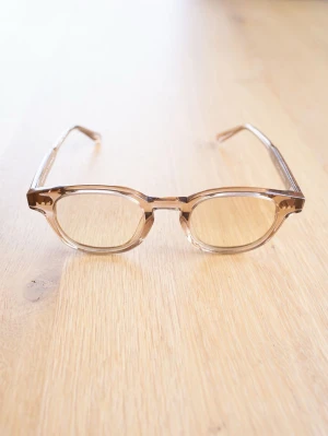 Chimi Solglasögon, Light Brown - Snygga glasögon med transparent beige båge och rundad form. Bågarna är designade i Stockholm och har en modern, stilren look som passar till många olika outfits. Tillverkade i plastmaterial för en lätt och bekväm känsla.