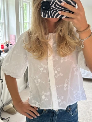 Vit blus med broderade blommor - Söt vit blus med puffiga korta ärmar och broderade blommor över hela tyget. Blusen har knäppning framtill och är gjord i ett tunt, lätt material. Aldrig använd med prislapp kvar!