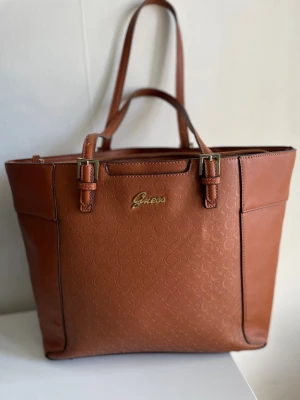 Fin Toteväska från Guess - Så fin brun Toteväska från Guess i läder🌟Väskan är använd men mycket fint skick! Plats för mycket saker. Lite skav på hörnet se sista bild men inget som stör. 