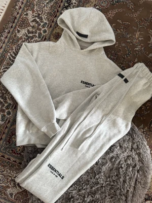 Essentials tracksuit - Snygga ljusgrå mjukisbyxor från Essentials Fear of God med tryckt logga på benet. Byxorna har resår i midjan och vid bensluten samt praktiska sidofickor. Perfekta för en avslappnad och trendig look Den har en liten fläck kom i Dm om du vill se den