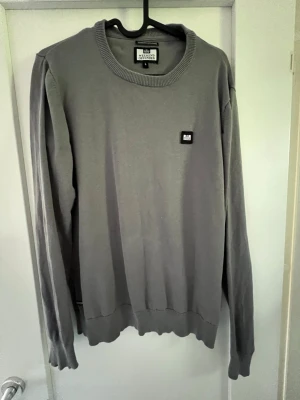 Grå tröja från Weekend offender - Snygg grå tröja från Weekend offender med rund halsringning och diskret logga på bröstet. Tröjan har långa ärmar och ribbade muddar vid ärmslut och nederkant. Perfekt till jeans eller chinos för en clean look. I bra skick. Är liten i storlek, skulle säga att den passar som S