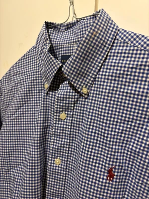 Blårutig skjorta från Ralph Lauren - Klassisk blårutig skjorta från Ralph Lauren med button-down krage och broderad logga på bröstet. Skjortan har långa ärmar, vita knappar och är tillverkad i mjuk bomull. Perfekt för en stilren och avslappnad look.