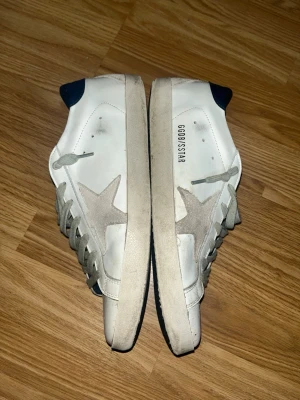 Vita Golden Goose sneakers med stjärna - Snygga vita sneakers från Golden Goose med grå mockastjärna på sidan och mörkblå detalj vid hälen. Skorna har grå skosnören och klassisk låg profil. Tillverkade i läder och mocka för en trendig och avslappnad look.