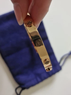 Guldigt armband med logodetaljer - Snyggt och stilrent armband i roséguld med Tory Burch klassiska logodetaljer runt om. Perfekt accessoar för att lyfta din outfit. Ytterst sparsamt använd och nypris över 2000 kr