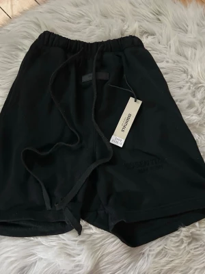 Svarta shorts från Essentials - Svarta shorts från Essentials med broderad logga på benet och bred resår i midjan. Shortsen har snörning i midjan för justerbar passform och är tillverkade i mjuk bomull, perfekta för avslappnade dagar. Klassisk och enkel design.