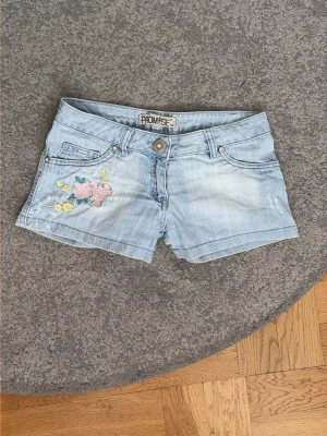 Ljusblå lågmidjade jeansshorts med broderi - Säljer ett par ljusblå lågmidjsde jeansshorts med broderade blommor i rosa och gult på ena fickan fram och bak. Shortsen har normal passform, låg midja, bälteshällor och klassisk femficksdesign. Perfekta för sommardagar och ger en fräsch look till din outfit. Vintage märket Promise. Storlek 36 men sitter mer som en 34. 