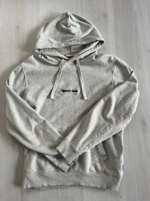 Ljusgrå hoodie från Jack & Jones - Snygg ljusgrå hoodie från Jack & Jones med huva och dragsnören. På bröstet finns en liten broderad text i svart där det står ”ORIGINAL STUDIOS”. Hoodien har en klassisk känguruficka framtill och ribbade muddar vid ärmslut och nederkant. Perfekt för en chill och avslappnad stil.
