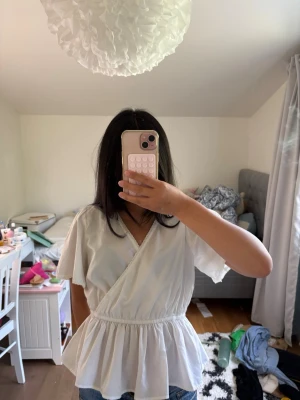 Vit omlottblus från Vero Moda - Säljer en söt vit omlottblus från Vero Moda med korta ärmar och volang nedtill. Blusen har en markerad midja och fina spetsdetaljer längs kanterna. Perfekt till jeans eller kjol för en fräsch look.
