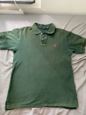  Polo Ralph Lauren - Klassisk mörkgrön pikétröja från Polo Ralph Lauren med röd broderad logga på bröstet. Tröjan har krage, knappar vid halsen och korta ärmar. Perfekt till en avslappnad men ändå stilren look.